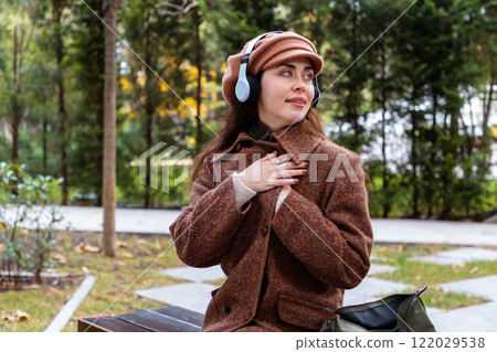 Mid shot of a beautiful dreamy young Caucasian woman in a hat and coat with headphones listening to music in the park Mid shot of a beautiful dreamy young Caucasian woman in a hat and coat with headphones listening to music in the park 122029538