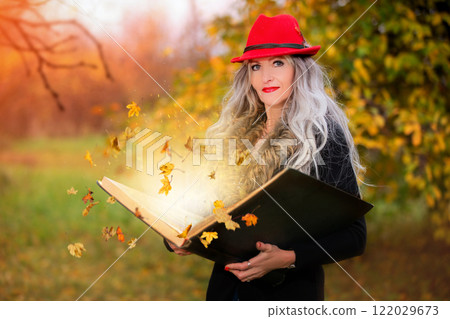 A beautiful woman with a book of magic spells with falling autumn leaves is conjuring 122029673