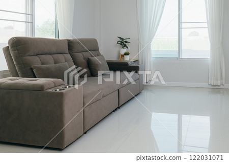 A modern gray sofa with two pillows and built-in drink holders, placed in a bright room with white curtains and floors. Minimalist living room interior with natural light and cozy ambiance. A modern gray sofa with two pillows and built-in drink holders, placed in a bright room with white curtains and floors. Minimalist living room interior with natural light and cozy ambiance. 122031071