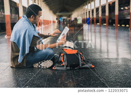 A young man traveler with digital tablet sitting on floor at train station choose where to travel. Concept of backpackers travel adventure by train. 122033071