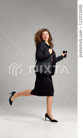 Confident young woman with curly hair, dressed formal outfit with black bag, jumping holding cup against minimalistic studio background. Confident young woman with curly hair, dressed formal outfit with black bag, jumping holding cup against minimalistic studio background. 122033606