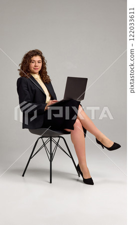 Confident young lady working in laptop, sitting in chair crossing legs against grey studio background. Freelancer. 122033611