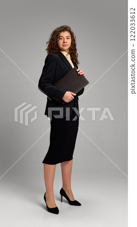 Portrait of young elegant, business lady dressed black formal outfit, holding laptop against minimalistic studio background. 122033612