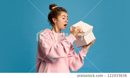 Young beautiful woman, dressed casual attire, opens box with curious and surprised expression against blue studio background. Young beautiful woman, dressed casual attire, opens box with curious and surprised expression against blue studio background. 122033636