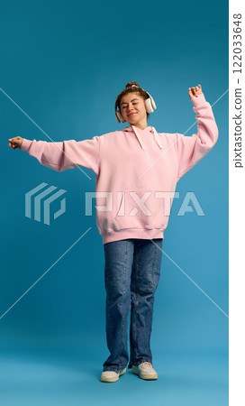 Happy young woman dressed pink hoodie, dancing raising hands while listening music in headphones against blue studio background. Happy young woman dressed pink hoodie, dancing raising hands while listening music in headphones against blue studio background. 122033648