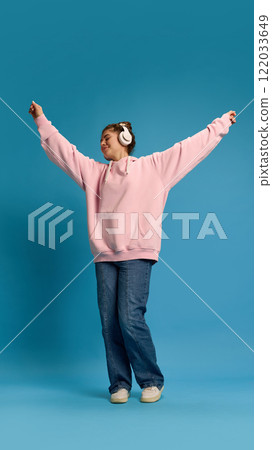 Overjoyed young woman dressed pink hoodie, dancing raising hands while listening music in headphones against blue studio background. Overjoyed young woman dressed pink hoodie, dancing raising hands while listening music in headphones against blue studio background. 122033649