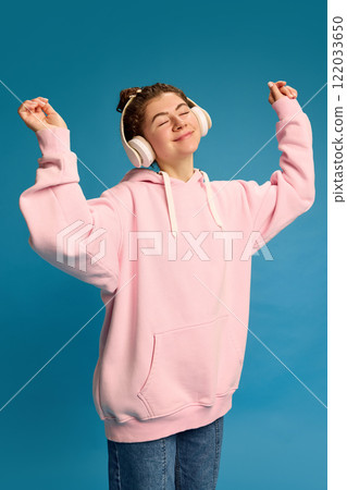 Positive, smiling young woman dressed pink hoodie, dancing raising hands while listening music in headphones against blue studio background. 122033650