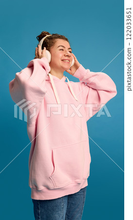 Positive, smiling young woman dressed pink hoodie, listening music in headphones with joyful expression against blue studio background. Positive, smiling young woman dressed pink hoodie, listening music in headphones with joyful expression against blue studio background. 122033651