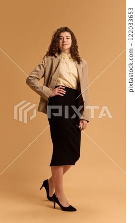 Proud and confident young businesswoman dressed formal attire posing holding hand on hip against sandy color studio background. 122033653