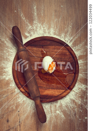 Cracked egg and rolling pin on a wooden board surrounded by flour during a cooking preparation 122034499