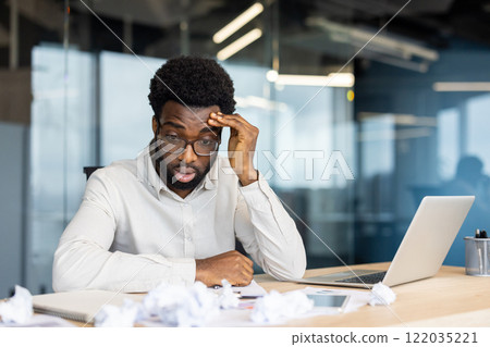 Disappointed and upset man doing paperwork inside office. Unsatisfied with financial reporting results sitting near crumpled pieces of papers. Businessman with laptop among documents. 122035221