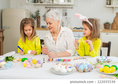 Happy Easter. Two little girls sisters twins grandmother painting eggs enjoying time together. Happy family grandma granddaughters child kids preparing for Easter. Spring Christian festival tradition 122035539
