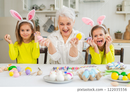 Happy Easter. Two little girls sisters twins grandmother painting eggs enjoying time together. Happy family grandma granddaughters child kids preparing for Easter. Spring Christian festival tradition 122035556