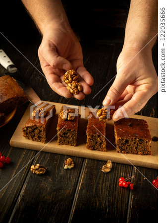 Hands carefully arrange walnuts on freshly baked squares of cake, showcasing a warm, inviting kitchen ambiance filled with rich aromas. The wooden board enhances the rustic charm of the moment 122035666