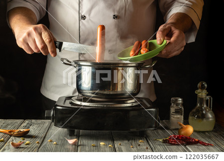 An experienced chef expertly adds a sausage to a steaming pot. The rustic ambience of the kitchen reflects a warm and inviting culinary experience, perfect for any food lover 122035667