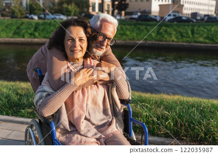 Senior woman in wheelchair walking with caregiver old man on road in park. Elderly family couple man supporting embracing woman in chair for people with disability outdoor. Rehabilitation Senior woman in wheelchair walking with caregiver old man on road in park. Elderly family couple man supporting embracing woman in chair for people with disability outdoor. Rehabilitation 122035987