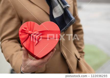 Happy man hold and gives heart-shaped red gift in beige coat on Valentine's Day. Close up. Copy space. Love concept for birthday or anniversary. Happy man hold and gives heart-shaped red gift in beige coat on Valentine's Day. Close up. Copy space. Love concept for birthday or anniversary. 122036293
