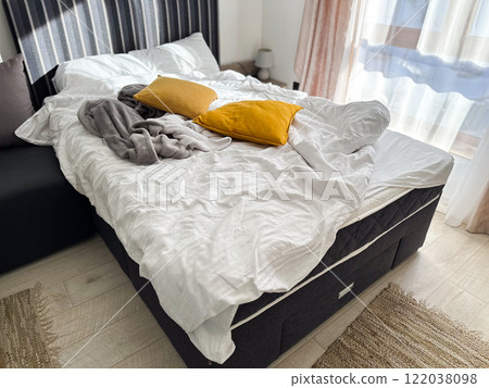 An unmade bed with a white blanket and mustard-yellow pillows in a bright, sunlit room. Mess at home concept An unmade bed with a white blanket and mustard-yellow pillows in a bright, sunlit room. Mess at home concept 122038098