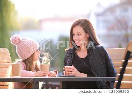 Young pretty woman having good time with her child daughter sitting at street cafe with hot drinks on sunny autumn day. Happiness in family relations concept Young pretty woman having good time with her child daughter sitting at street cafe with hot drinks on sunny autumn day. Happiness in family relations concept 122039230