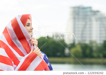Young happy refugee woman with USA national flag on head and shoulders. Positive muslim girl traveling in United States. 122039242