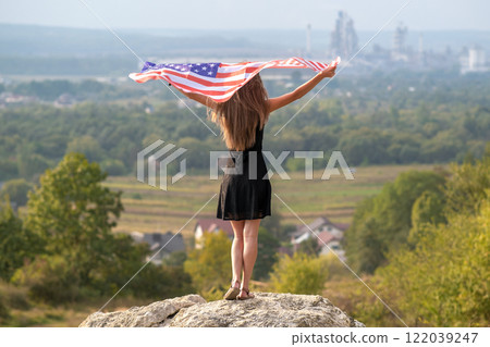 Young happy american woman with long hair raising up waving on wind USA national flag in her hands relaxing outdoors enjoying warm summer day. 122039247