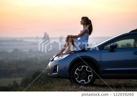 Yong happy female driver resting on hood of her car enjoying sunset view of summer nature. Travel destinations and recreation concept. Yong happy female driver resting on hood of her car enjoying sunset view of summer nature. Travel destinations and recreation concept. 122039260