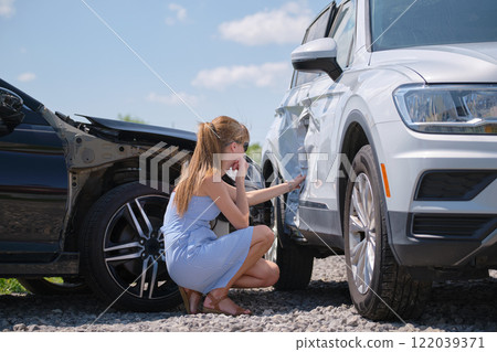 Sad young woman driver sitting near her smashed car looking shocked on crashed vehicles in road accident Sad young woman driver sitting near her smashed car looking shocked on crashed vehicles in road accident 122039371