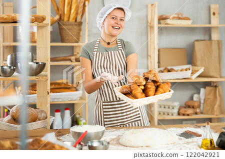 Young woman baker are standing near product range in bakery kitchen with basket of croissants. Young woman baker are standing near product range in bakery kitchen with basket of croissants. 122042921