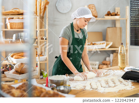 Young baker girl is standing near work table in kitchen, working with dough Young baker girl is standing near work table in kitchen, working with dough 122042947