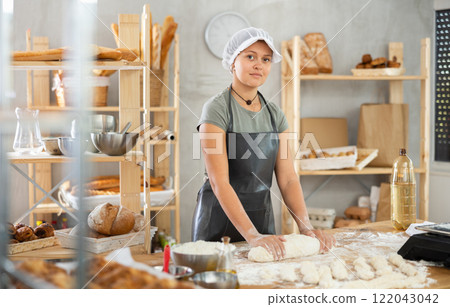 Young baker girl is standing near work table in kitchen, working with dough Young baker girl is standing near work table in kitchen, working with dough 122043042
