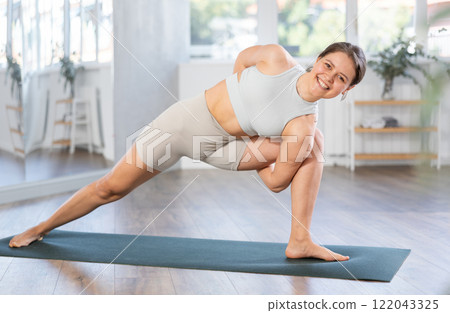 Sporty energetic female teacher doing exercises on pilates mat and demonstrating variation of warrior pose of yoga at gym. Fitness concept 122043325