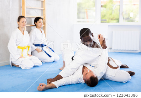Judo or jiu-jitsu classes - two men practicing grabbing and throwing on sports mats under the guidance of trainer 122043386