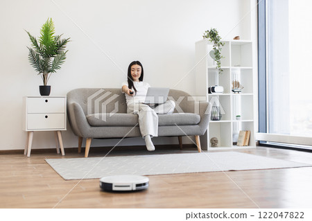 Asian woman sitting on couch using laptop and remote while robot vacuum cleans floor. Cozy modern living room with bright decor. 122047822