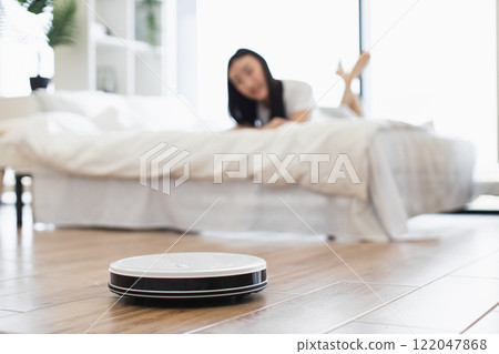 Asian woman enjoying time in bedroom while robot vacuum cleaner efficiently cleans floor. Modern technology aiding household chores, offering convenience and relaxation. 122047868