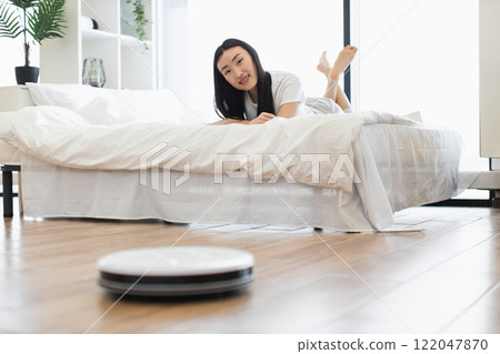Asian woman relaxing on bed while robot vacuum cleaner cleans modern minimalistic bedroom. Concept of technology, cleanliness, and comfort in daily life. 122047870