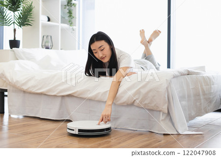 Asian woman relaxing on bed while controlling robot vacuum cleaner. Concept of modern technology, convenience, smart home lifestyle. Young adult enjoying clean and tidy living space. 122047908
