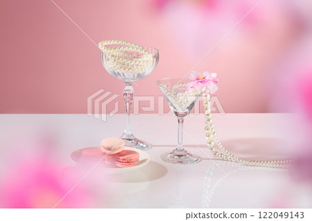 A plate of macarons, a few pearl-string glasses, pink flowers, and an empty space for product displays are shown in front. The background is pink. A party-themed advertisement for cosmetics. A plate of macarons, a few pearl-string glasses, pink flowers, and an empty space for product displays are shown in front. The background is pink. A party-themed advertisement for cosmetics. 122049143