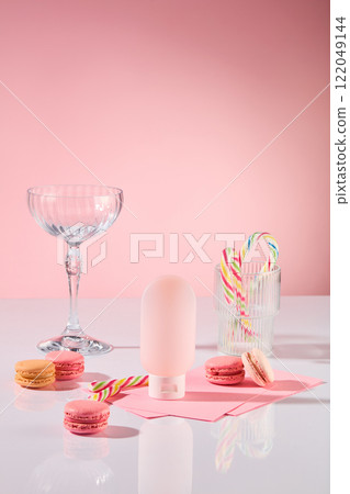 An advertising product photo with a close-up angle. Party table theme featuring macarons, candy canes in a glass, and a pink cosmetic bottle mockup on a white surface against a pink backdrop. 122049144