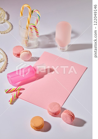 An overhead view of a cosmetics ad. The dessert table motif includes a mockup of a pink cosmetic bottle on a white surface, next to pink macarons, pearls, candy canes, a clear glass, and pink paper. 122049146