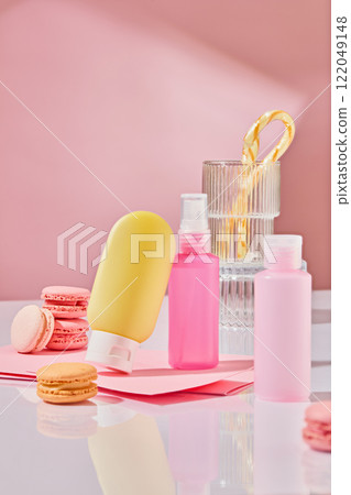 A frontal shot shows cosmetic bottle mockups and macarons; a glass holds a candy cane arranged on a white surface against a pink backdrop. The theme is a dessert table for a cosmetics ad. 122049148