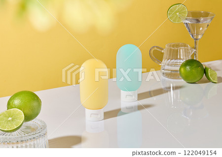 Side angle shot of two squeeze bottles positioned centrally on a white surface. Limes, a martini glass, and a glass pitcher arranged against a yellow background. The fruit theme for cosmetics ads. 122049154