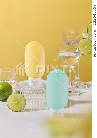 Close-up shot of two squeezable bottle mockups perched on a glass pedestal. Limes, some martini glasses, and a glass water pitcher are arranged around. The yellow backdrop, perfect for a cosmetics ad. 122049155