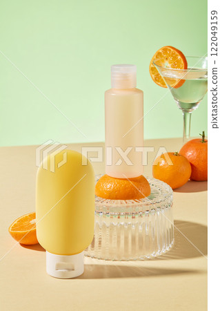 A cosmetics advertisement features a pale orange bottle on an orange placed on a glass podium, a yellow bottle on a beige surface, oranges, and a cocktail glass, amidst a green backdrop. 122049159