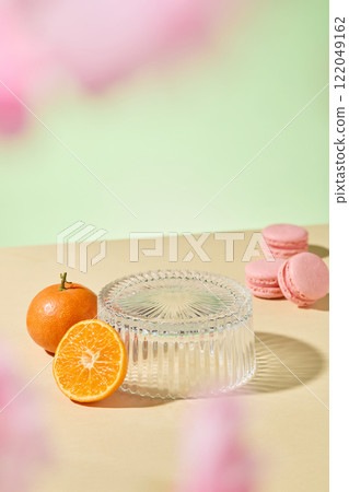 Some pink macarons and some orange slices placed next to an empty glass podium for present product. Against a green background with a beige surface. Overhead shot. An advertisement photo for cosmetics 122049162