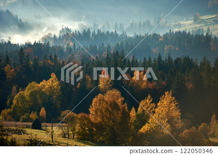 Autumn foliage in vibrant hues of gold, red, and orange blankets the forested hills, with mist rising gently in distance. Haystack in sunlit meadow, adding rustic charm to serene landscape. 122050346