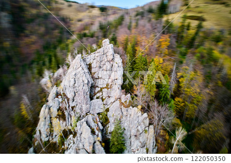 Dynamic aerial shot captures the jagged peaks of rocks adorned with flag. Blurred, colorful forest below adds sense of motion and depth, emphasizing dramatic and rugged nature of rocky outcrop. 122050350