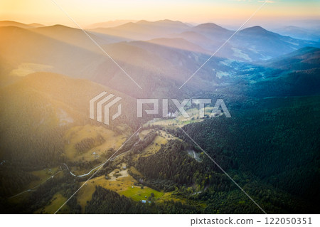 Aerial view of valley nestled between forested mountains, bathed in golden light from setting sun. Landscape features winding road, scattered patches of farmland, creating serene and picturesque scene 122050351