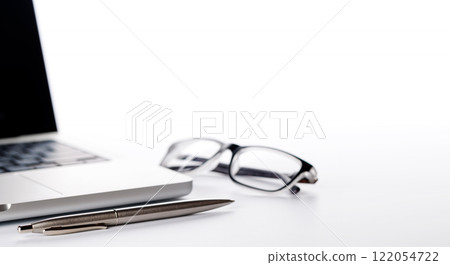 Closeup of an office table featuring a sleek laptop, a pen, and reading glasses on a clean white surface 122054722