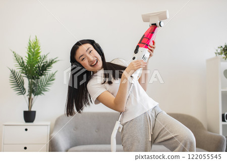 Asian young woman enjoying cleaning with headphones and vacuum. Happy and playful atmosphere in modern living room setting. Asian young woman enjoying cleaning with headphones and vacuum. Happy and playful atmosphere in modern living room setting. 122055545