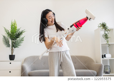 Asian young woman wearing headphones joyfully cleaning living room with vacuum, embracing fun in household chores. Asian young woman wearing headphones joyfully cleaning living room with vacuum, embracing fun in household chores. 122055575